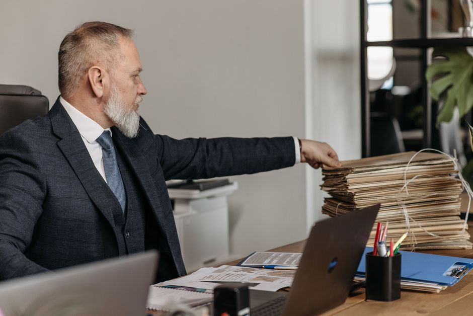 A mature man with a grey beard, wearing a dark checkered suit and blue tie, seated at a desk in an office. He is reaching out with his right hand to touch a large stack of manila folders tied with whi