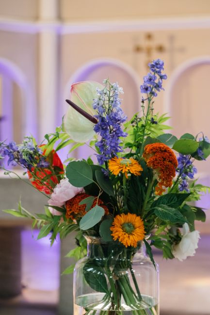 A vibrant floral arrangement in a clear glass vase features a variety of fresh flowers including bright orange marigolds and yellow gerbera daisies, complemented by purple and blue delphiniums and del