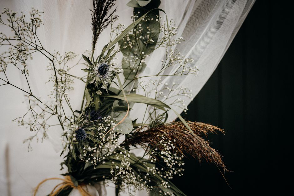 A close-up of a floral arrangement featuring a variety of dried and fresh flowers. The bouquet includes blue thistle blooms, small white baby's breath, delicate white gypsophila, and long, slender gre