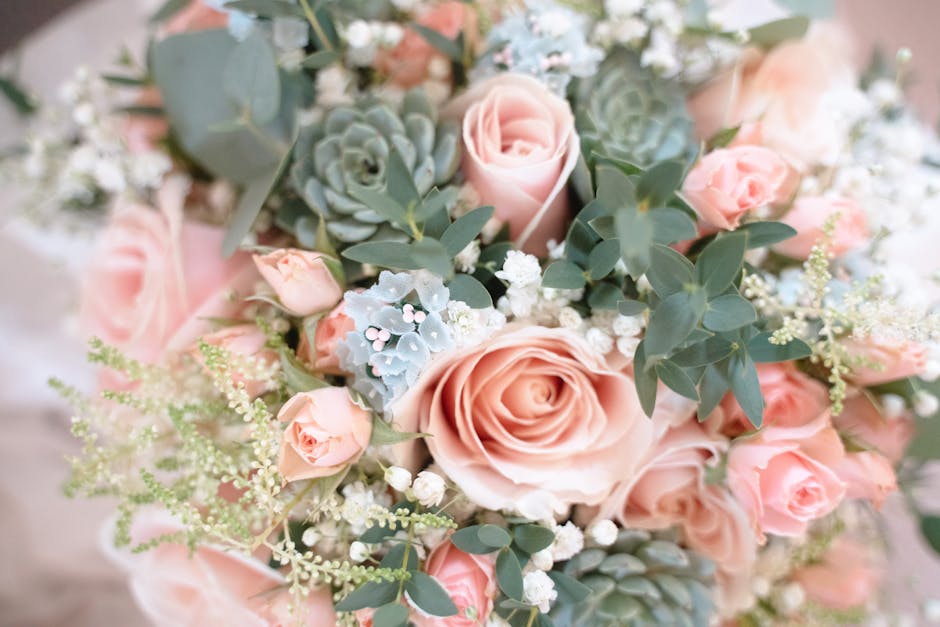 A close-up view of a floral bouquet featuring soft pink roses, small pink spray roses, pale blue succulents, and delicate white baby's breath, all arranged with lush green eucalyptus leaves. The bouqu