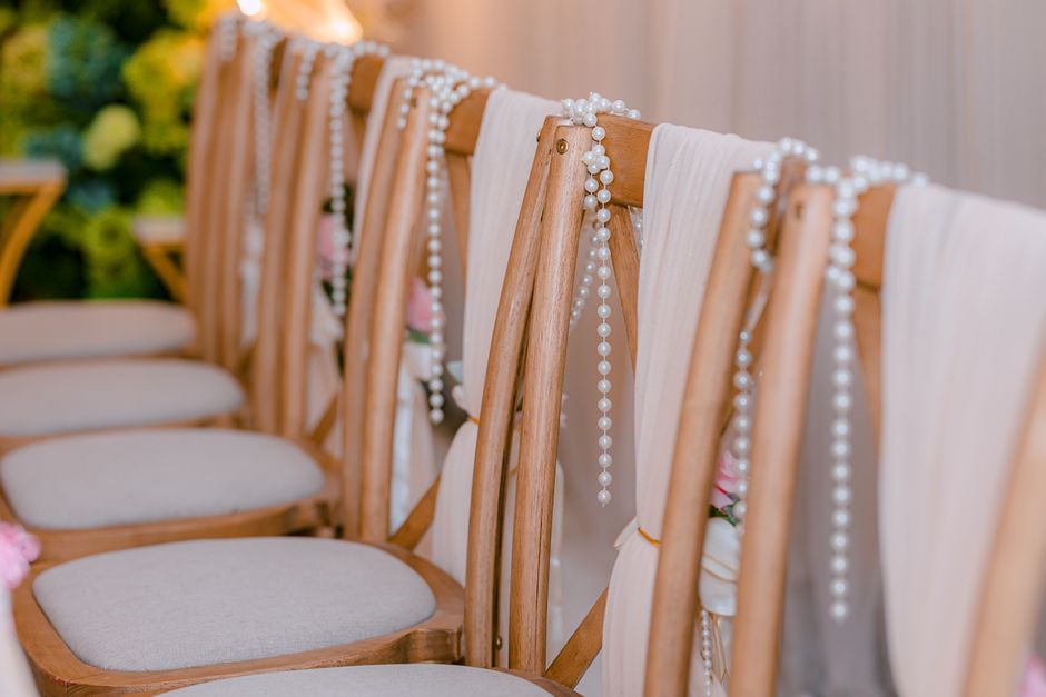 A row of elegant wooden chairs with cream-colored upholstered seats, decorated with delicate strands of white pearl beads draped over the backrests. Small floral arrangements featuring soft pink roses